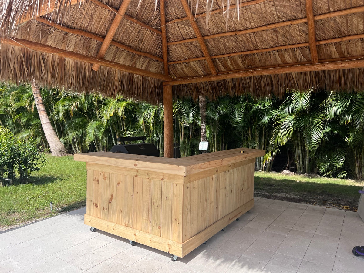 Natural wood L-shaped bar beneath a thatched roof surrounded by palms